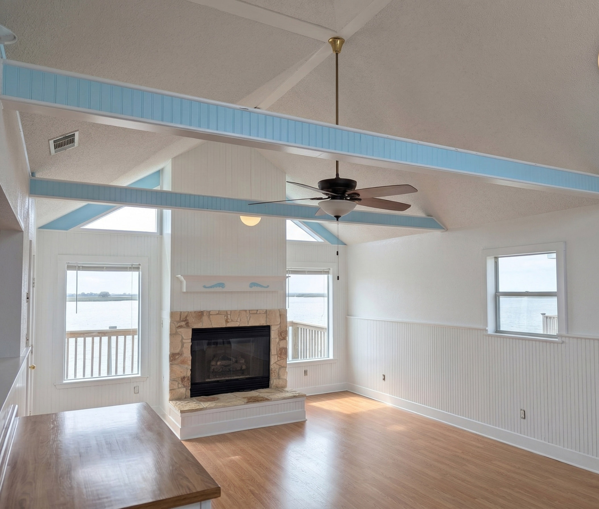 Bright coastal-style living room interior with vaulted ceilings, white wainscoting, light blue accent beams, a stone fireplace, and water views through large windows